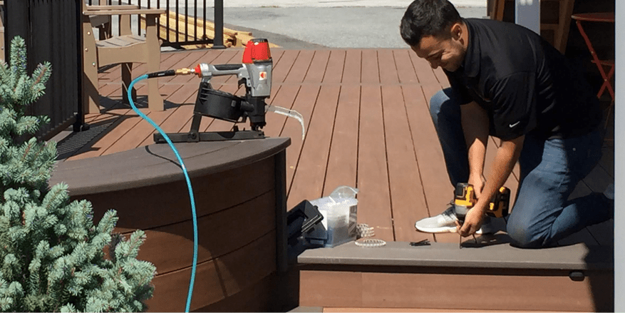 A man with brown hair in jeans and a black Nike polo is drilling screws into a new brown wooden deck near the stairs. There are building materials, pine trees, deck furniture, and power tools surrounding him.